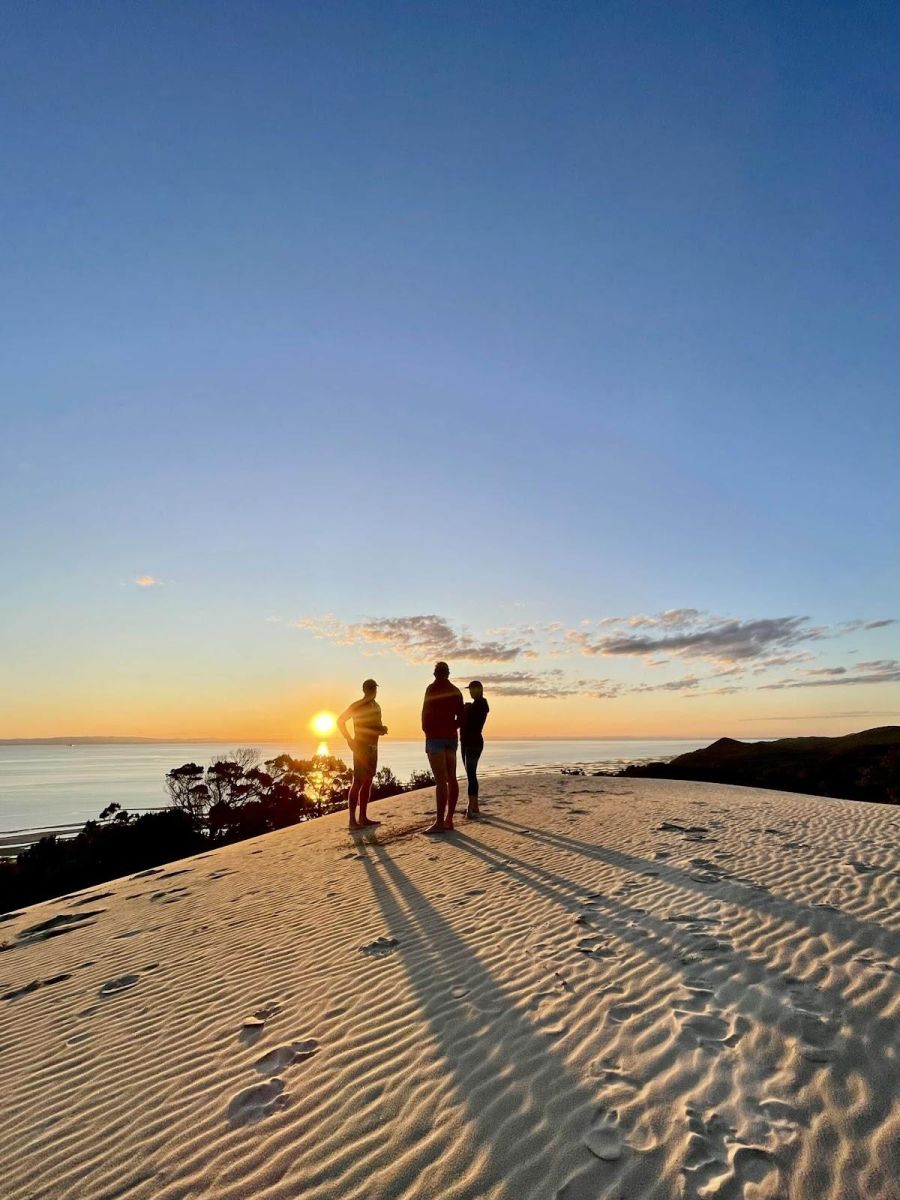 Sillouette of 3 people standing on top of a sand dune look out with the sun setting