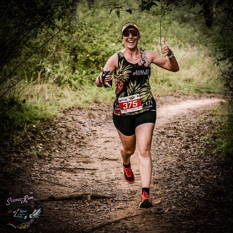 Amelia running on a trail wearing a Wild Earth race shirt and smiling